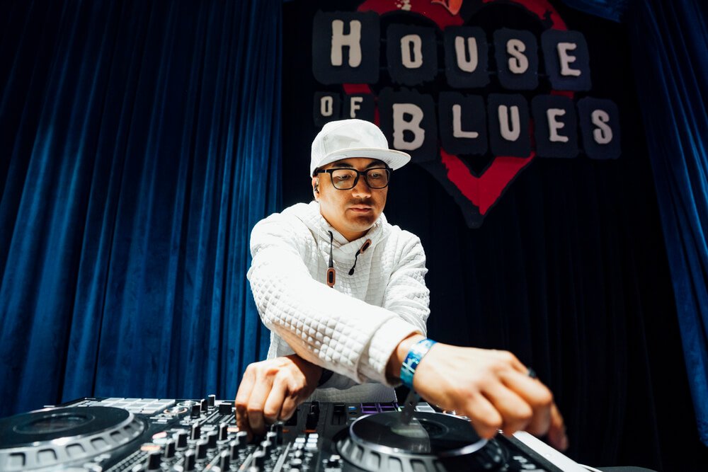 A DJ in a white outfit and cap adjusting equipment on stage at the House of Blues, with the venue's logo prominently displayed in the background, organized by emc3, a global events agency