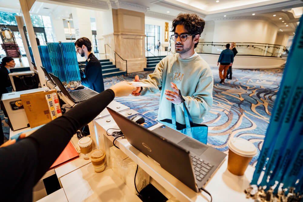 A conference attendee receiving a badge at the registration desk, with staff assisting and event lanyards visible in the background