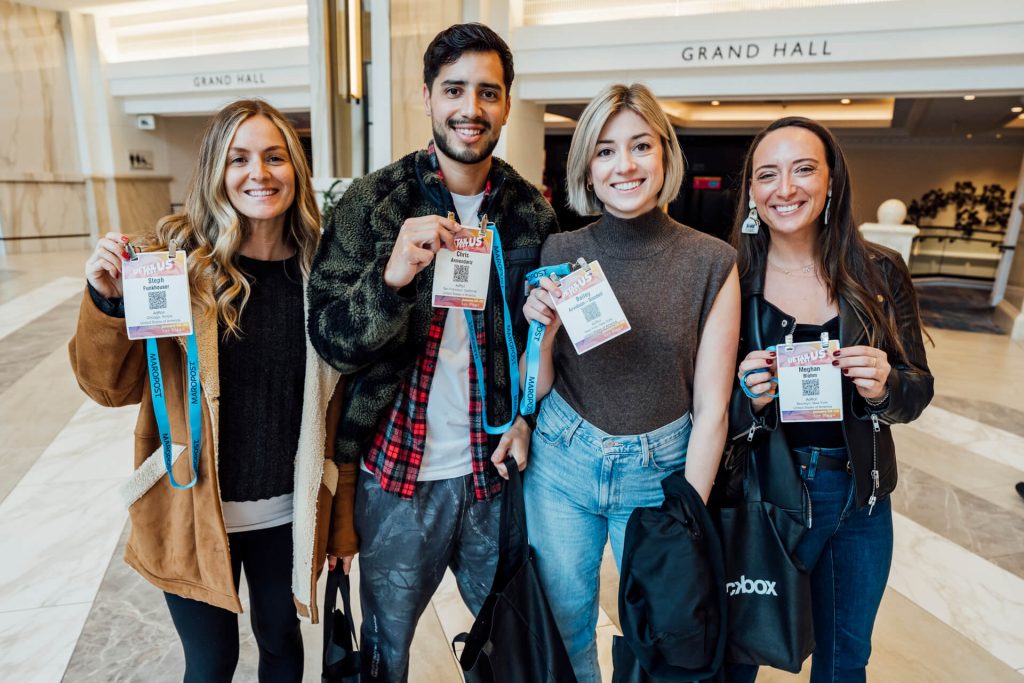 A group of four attendees at a conference proudly displaying their event badges and smiling for the camera in a well-lit lobby area