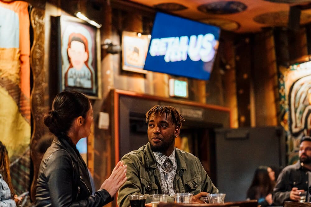 A man and a woman engaged in a conversation at a bar-like setting, with colorful artwork and a screen displaying 'Retail Us' in the background, organised by emc3, a global events agency