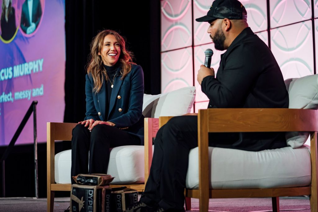 A panel discussion at a conference, featuring a female speaker smiling and engaged in conversation with a male interviewer on stage