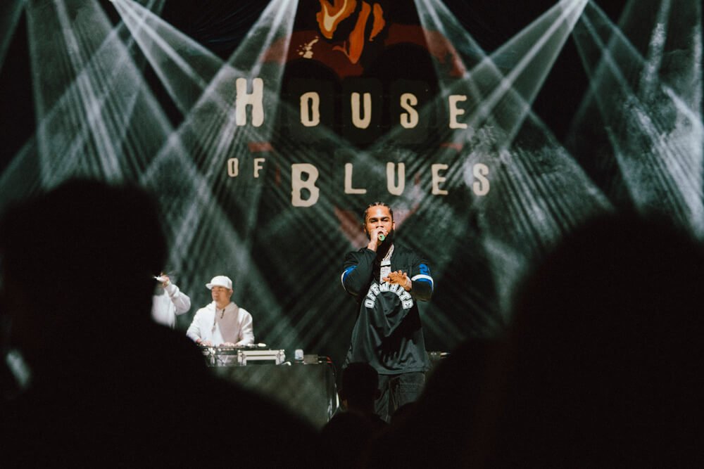 A performer on stage at the House of Blues, engaging with the audience, with a DJ in the background and dramatic lighting effects, organised by emc3, a global events agency