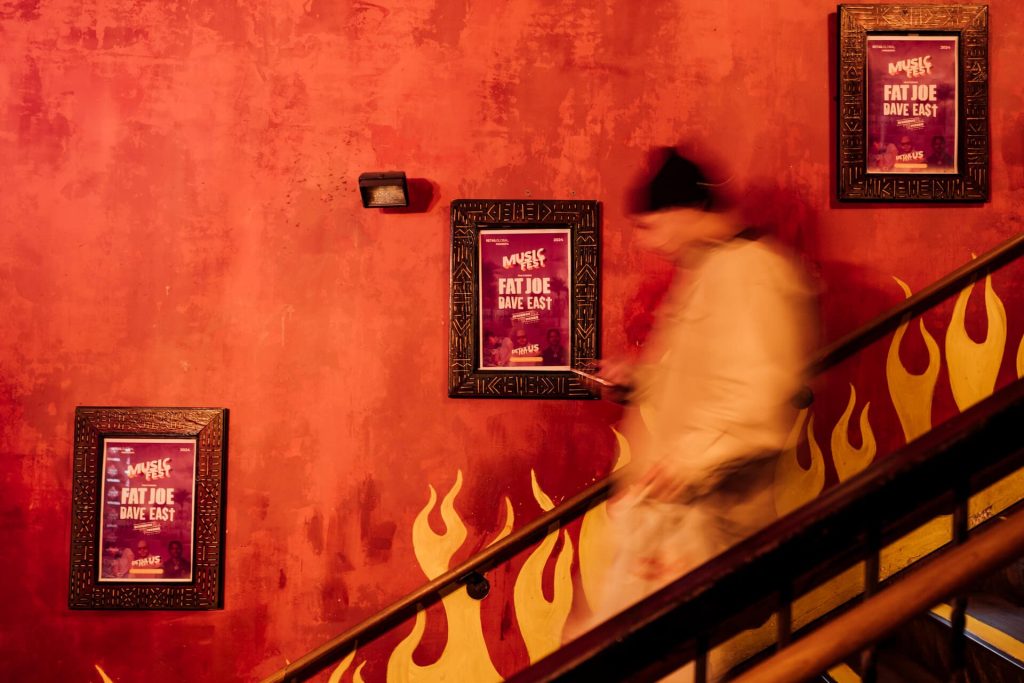 An individual descending a staircase with vibrant red walls adorned with promotional posters for a music fest featuring Fat Joe and Dave East