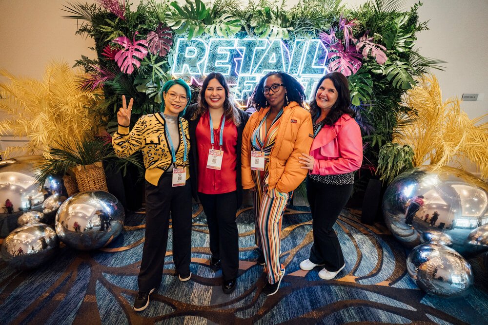Four diverse women posing in front of a 'Retail Fest' neon sign, with vibrant decorations and a cheerful atmosphere, organised by emc3, a global events agency