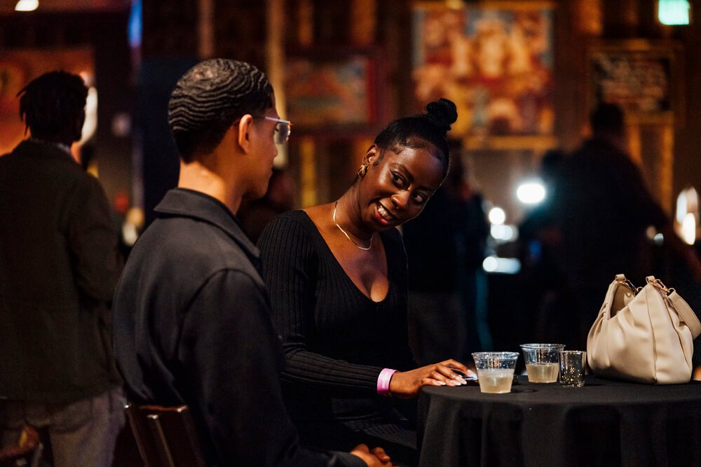 Two people enjoying a conversation at a dimly lit event, sitting at a table with drinks, and an audience in the background, organised by emc3, a global events agency