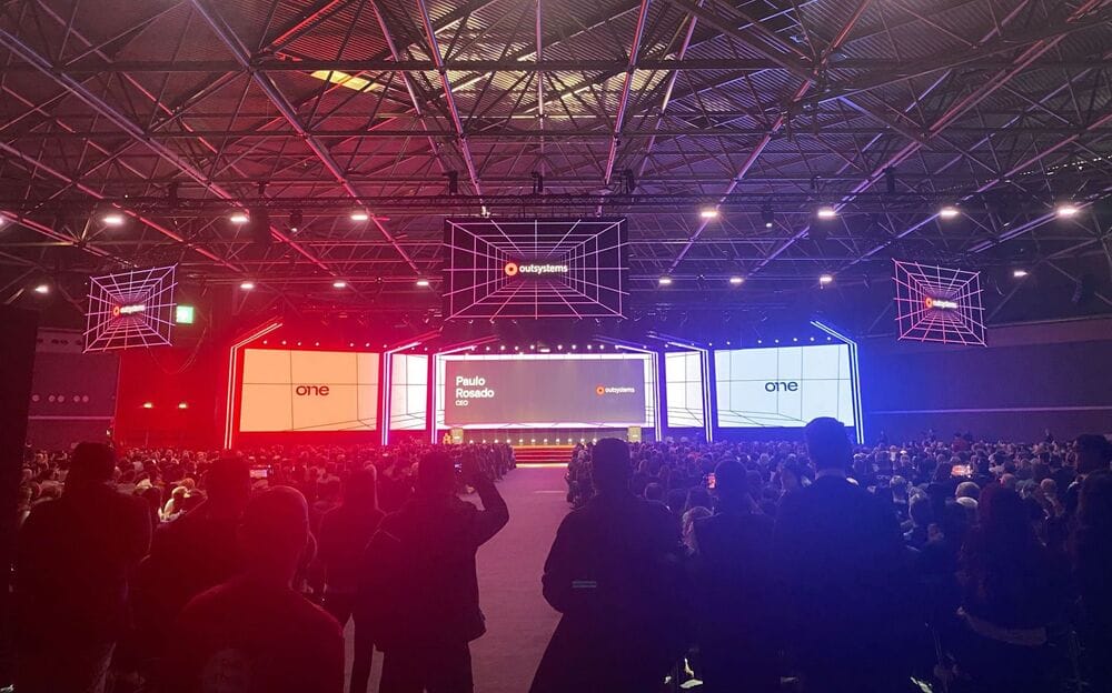 A large audience at a tech conference with a brightly lit stage and screens displaying OutSystems